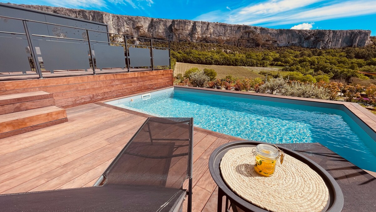 The image captures a serene pool area with a refreshing, clear water pool and wooden decking. A comfortable lounger rests beside the pool, accompanied by a small table featuring a jar. A panoramic view of the cliff and greenery is visible in the background.