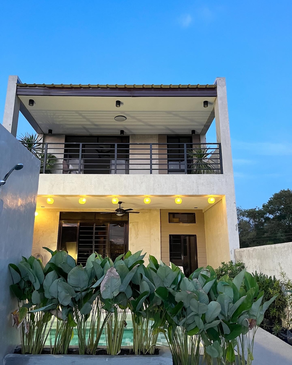 The two-story vacation home features a modern design with a balcony and large glass doors. Lush green plants line the front, and warm lights illuminate the space, creating a welcoming ambiance as the evening sky deepens into blue.