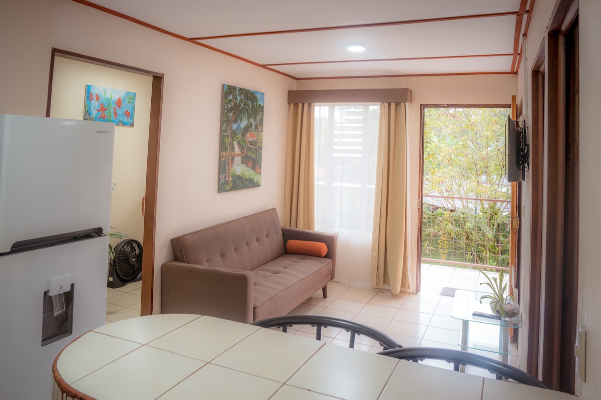 A bright living area is featured with a comfortable sofa and a flat-screen television mounted on the wall. Large windows allow natural light to fill the space, while colorful artwork decorates the walls. A dining area with a tiled table is visible in the foreground.
