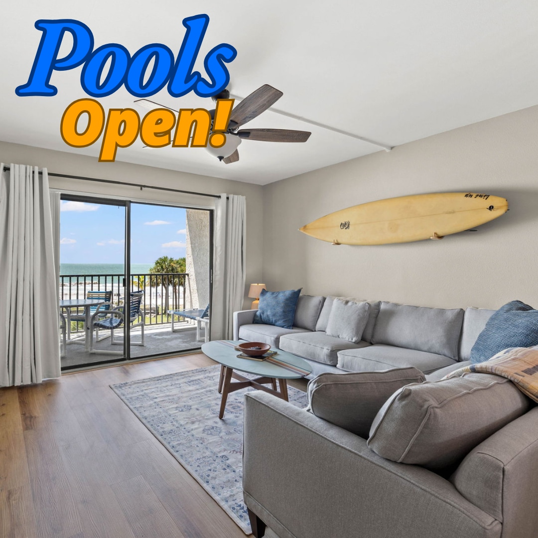 A cozy living area features a comfortable gray sofa with decorative pillows and a light wood coffee table. A surfboard enhances the beach-inspired decor. Natural light filters through sliding glass doors, which open to a patio with views of the beach and pool area.