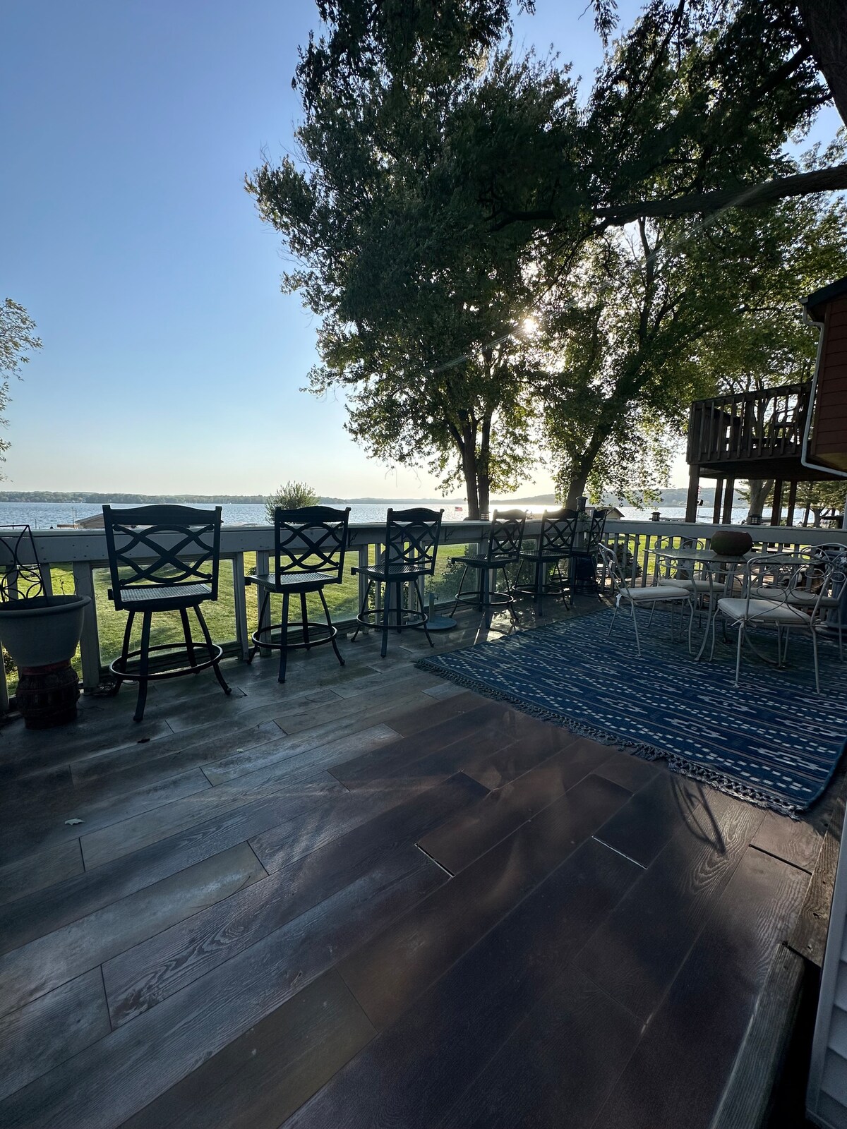 An outdoor deck area is visible, featuring bar-height seating aligned along a railing with views of the lake. The floor combines dark wood tones, complemented by a blue patterned rug. Lush greenery and sunlight create a serene atmosphere for relaxation.