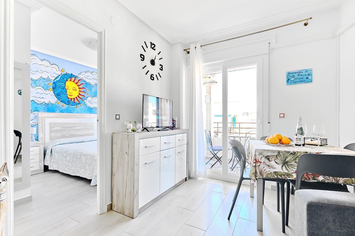 A bright and airy living space features a dining table with chairs and a sideboard, complemented by a wall adorned with a colorful mural of a sun and clouds. A window extends to a balcony, allowing natural light to fill the area.