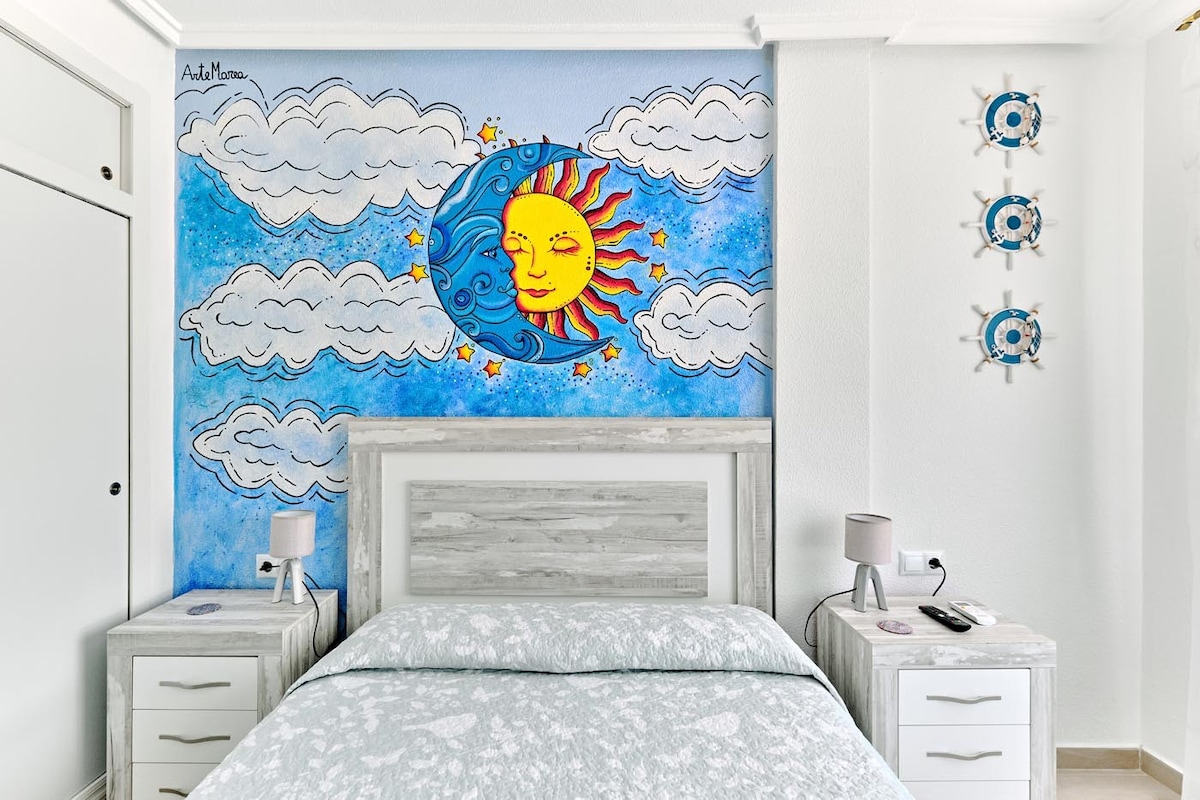 A bright and inviting bedroom features a large mural of a sun surrounded by clouds on a blue backdrop. The spacious bed is framed by nightstands with lamps on either side, while minimalistic decor adds to the overall ambiance.