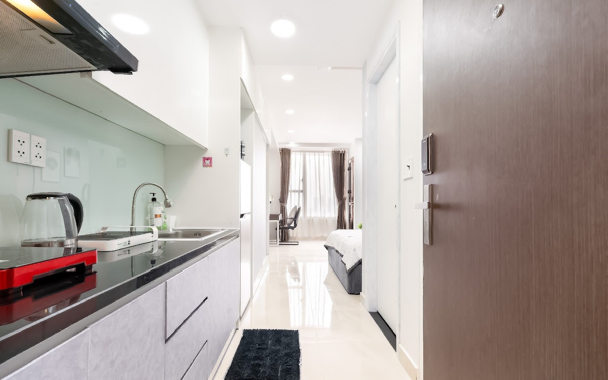 A well-equipped kitchenette is visible, featuring a sleek design with modern appliances and ample counter space. The view extends into a bright living area, where natural light flows through large windows. A cozy bed is positioned in the background, along with a comfortable chair.