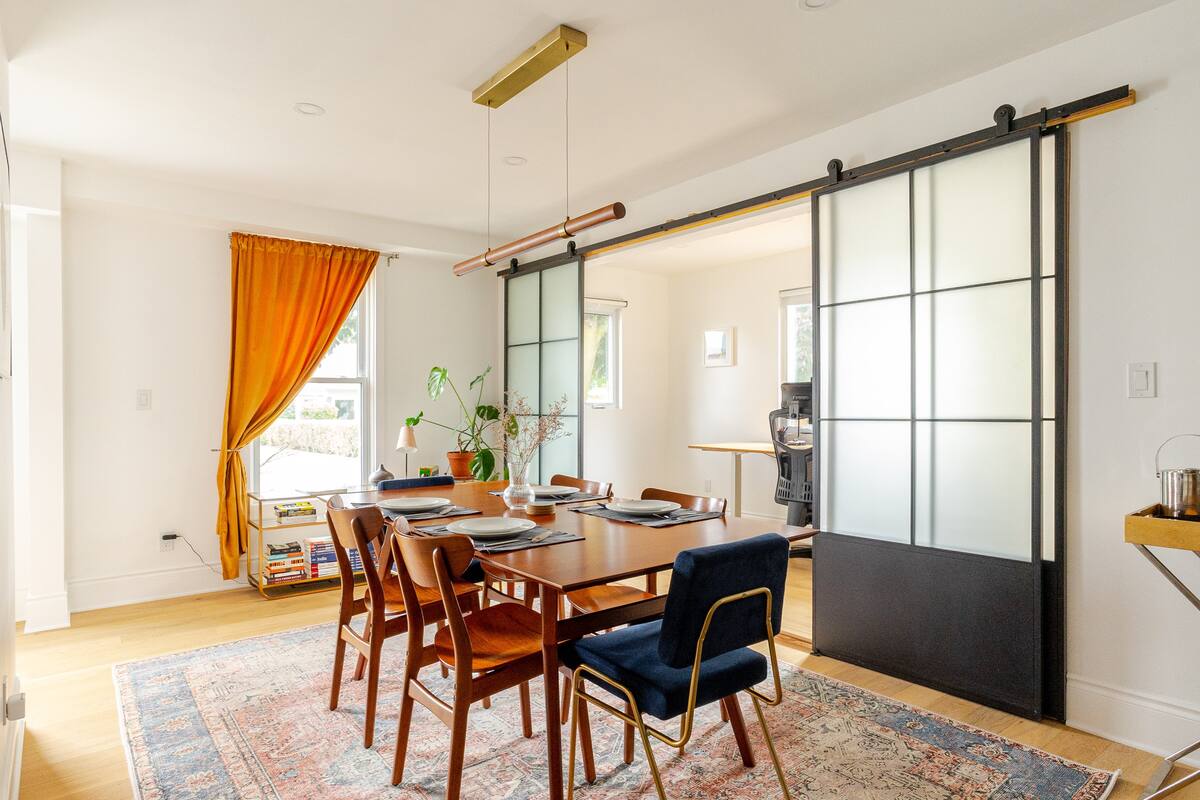 A dining area features a wooden table surrounded by stylish chairs. Natural light brightens the space, complemented by a warm orange curtain. Two sliding glass doors separate this area from an office space, providing a modern touch to the room's design.