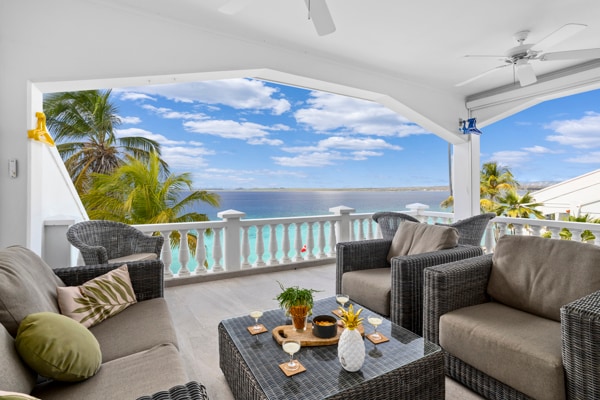 A spacious outdoor seating area is presented with comfortable wicker chairs and a coffee table adorned with decorative items. The stunning ocean view is framed by an expansive open space, featuring a bright sky and palm trees in the background.