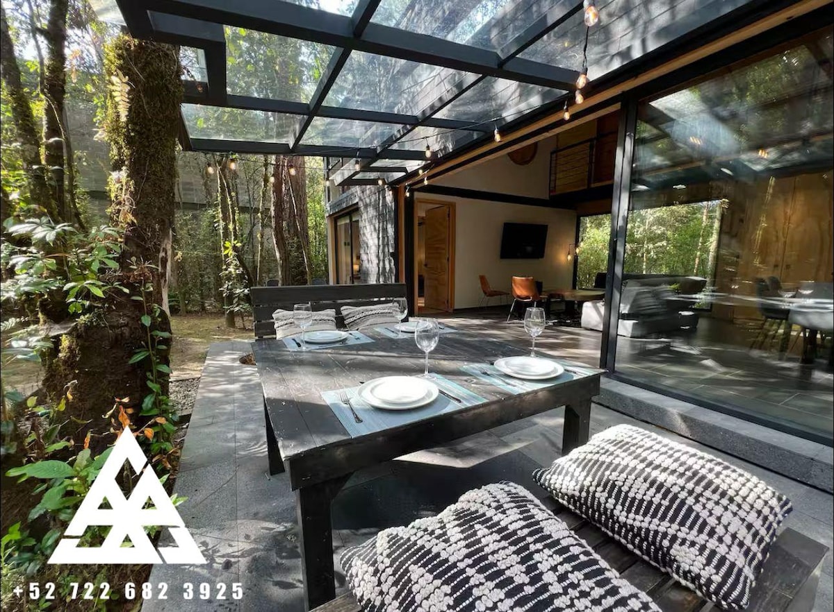 An outdoor dining area is presented with a wooden table and decorative pillows arranged on a stone surface. Glass panels create a feeling of openness to the surrounding forest. Soft lighting strings hang overhead, enhancing the connection between the interior and natural environment.