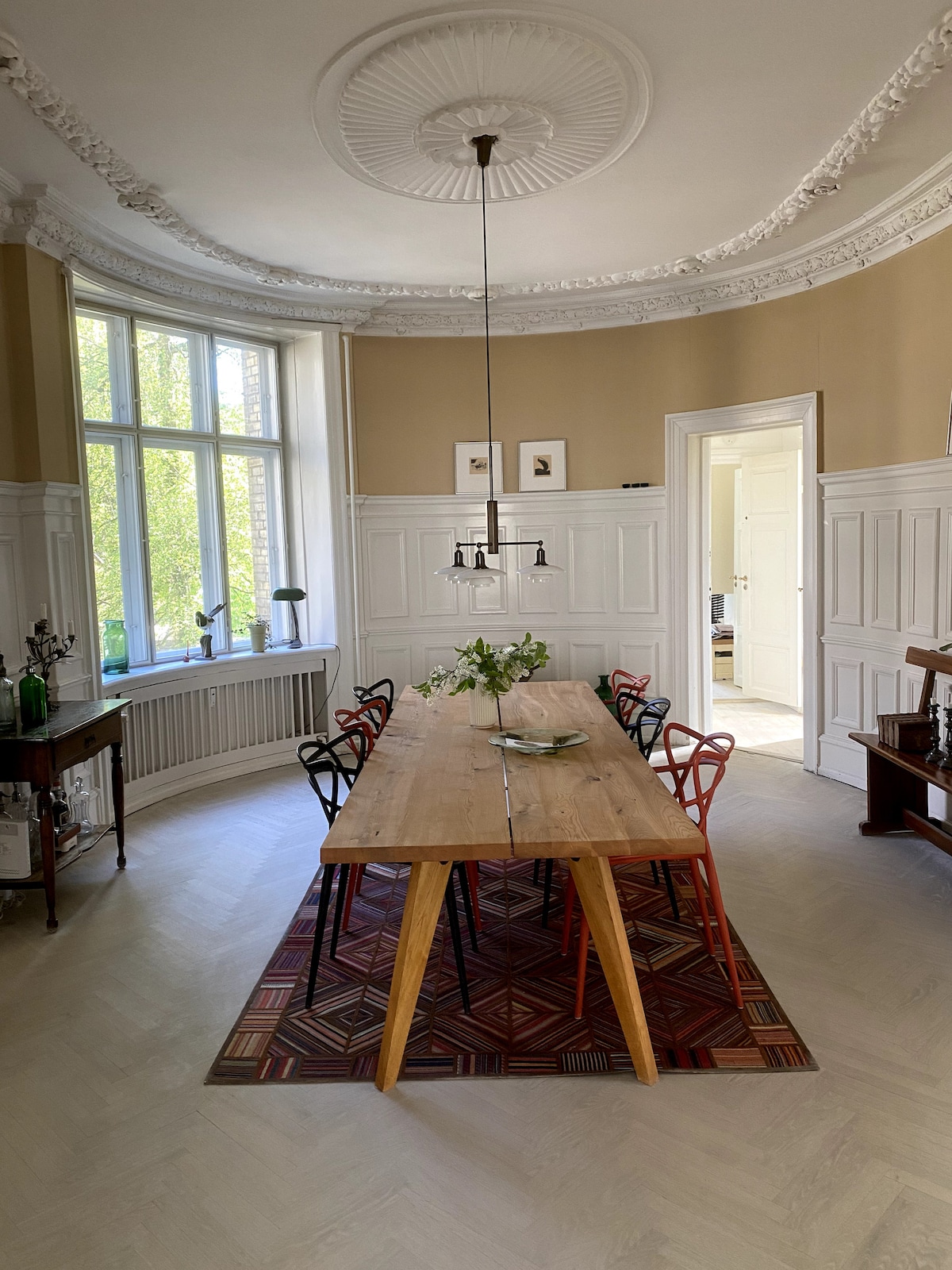 A well-lit dining area showcases a long, wooden table surrounded by colorful chairs. The room features large windows allowing natural light to flood in, highlighting the intricate ceiling details. A patterned rug is placed beneath the table, with a small floral arrangement serving as a centerpiece.