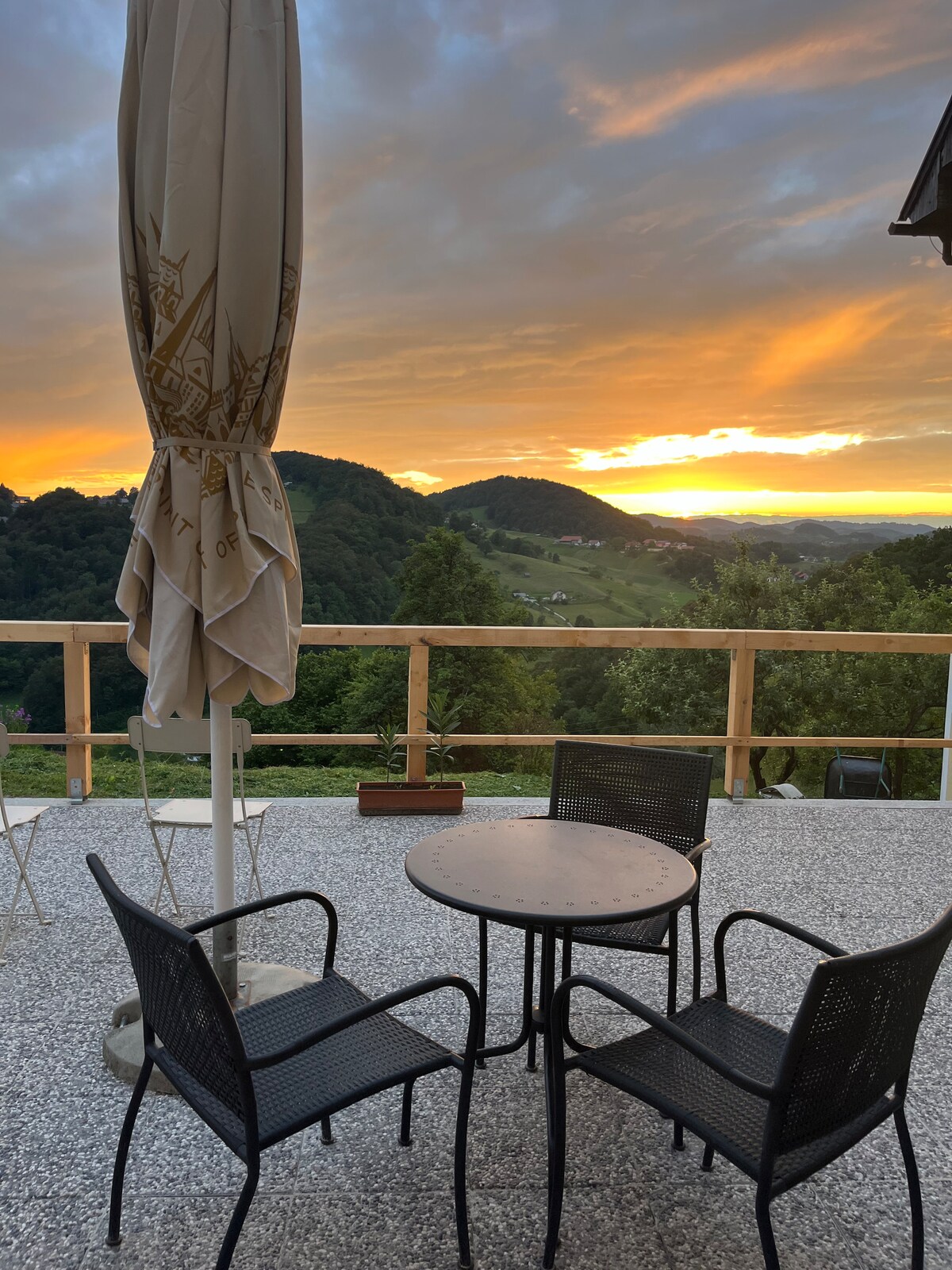 An outdoor seating area features a round table surrounded by four black chairs, positioned on a textured surface. A large umbrella provides shade. In the background, a sunset casts warm colors over rolling hills and greenery.