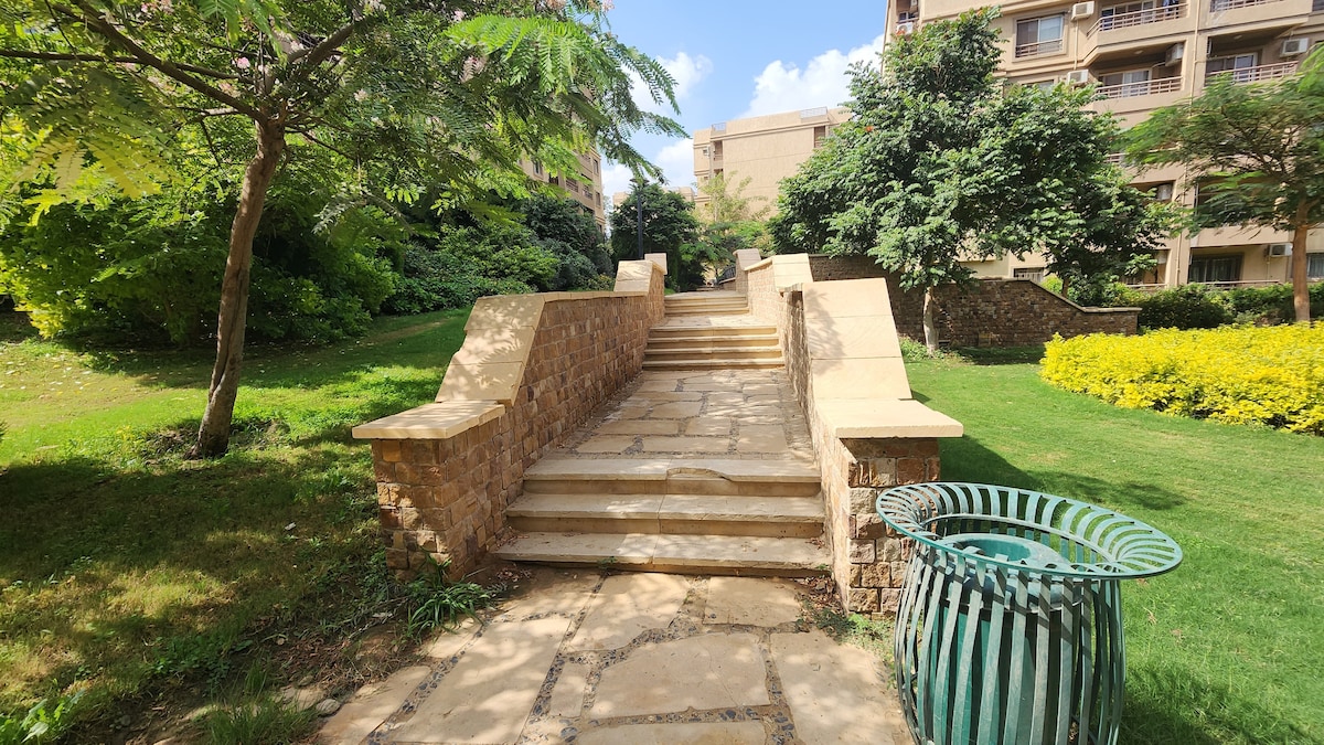 A landscaped area is visible, featuring a stone pathway leading up a set of stairs. Lush greenery surrounds the pathway, with trees and flowering plants complementing the scenery. A green waste bin is positioned nearby, enhancing the tidy appearance of the outdoor space.