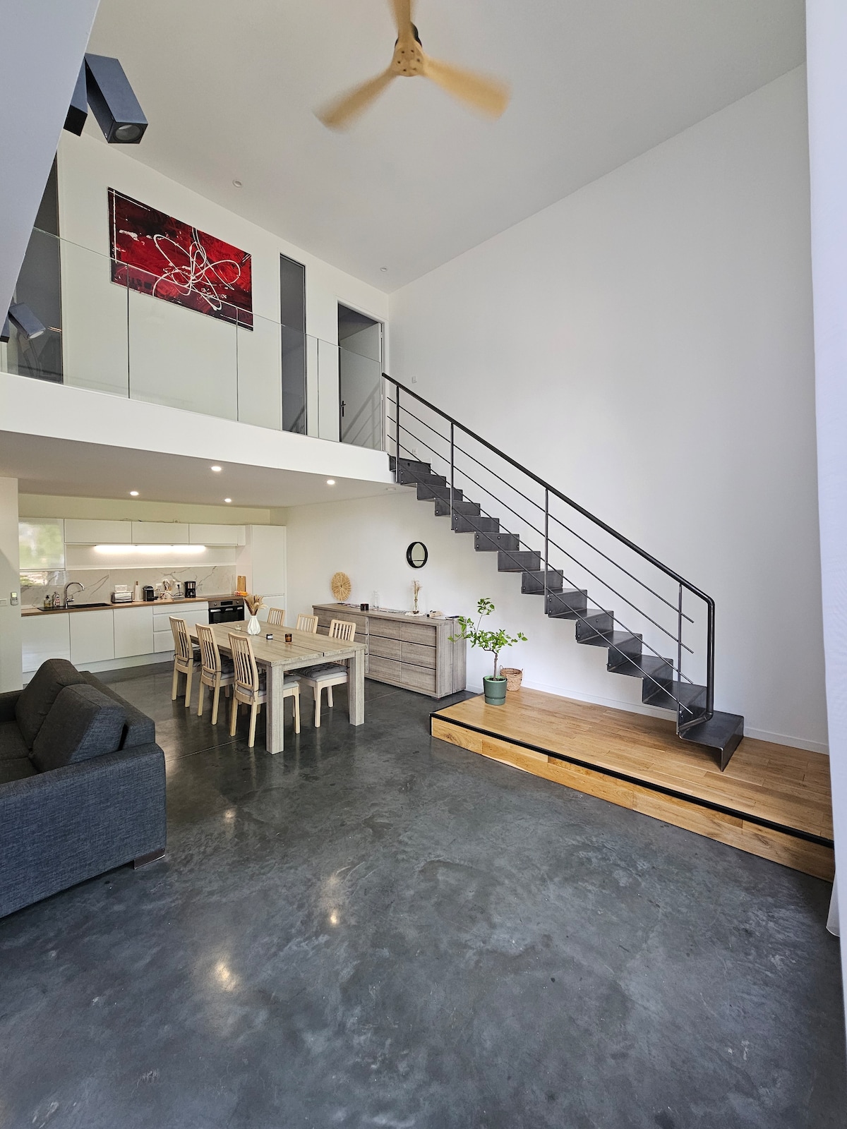 An airy loft space features a modern, open-concept living area with a sleek staircase leading to a second level. A dining table with light-colored chairs sits beside a comfortable sofa, surrounded by large windows that flood the area with natural light. Minimalist decor enhances the ambiance.