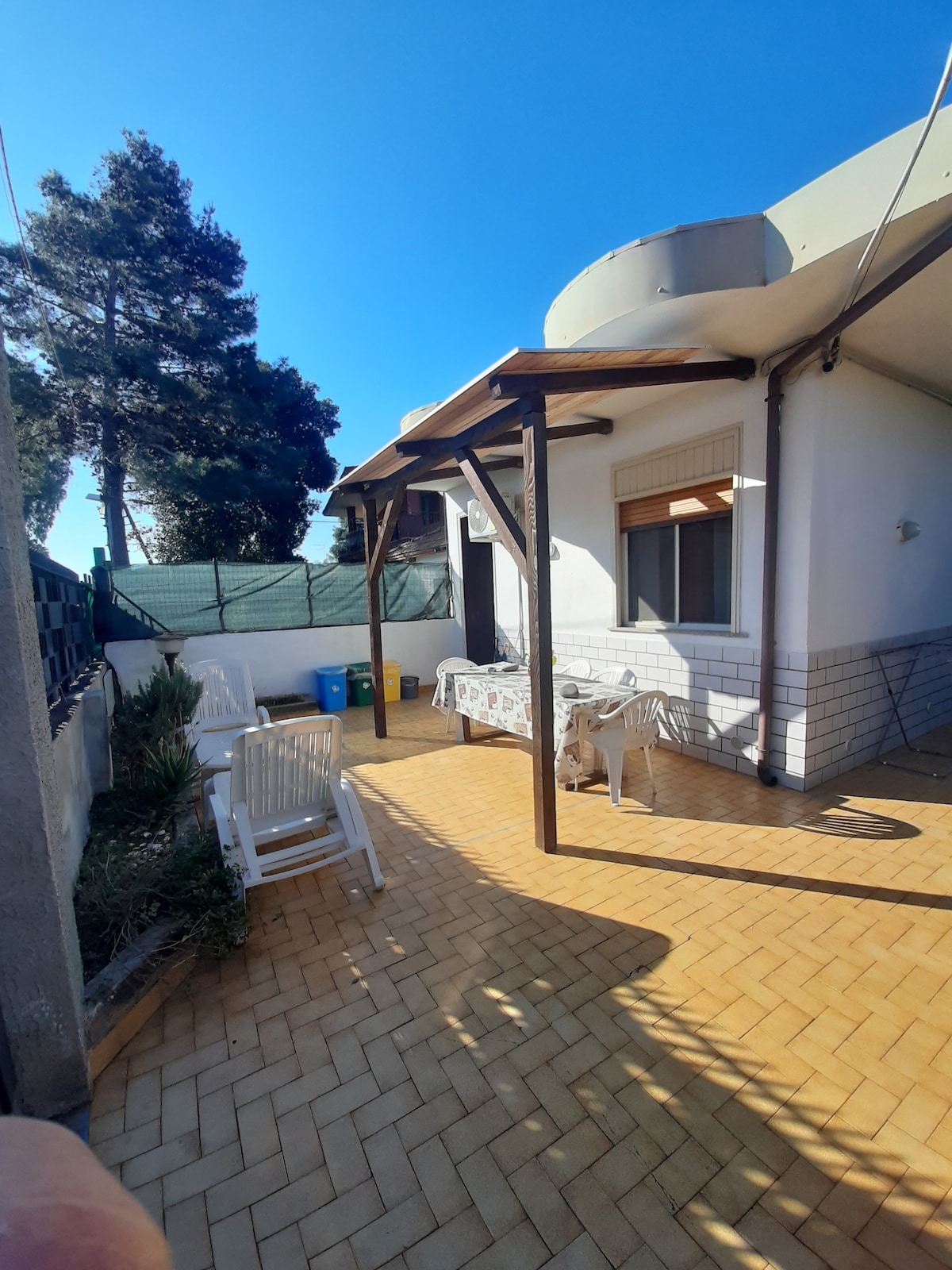 A sunlit patio area is shown, featuring a seating arrangement with white chairs and a table. Lush greenery is visible nearby, along with a privacy fence. Colorful waste bins are positioned discreetly in the background. The clear blue sky enhances the outdoor space.