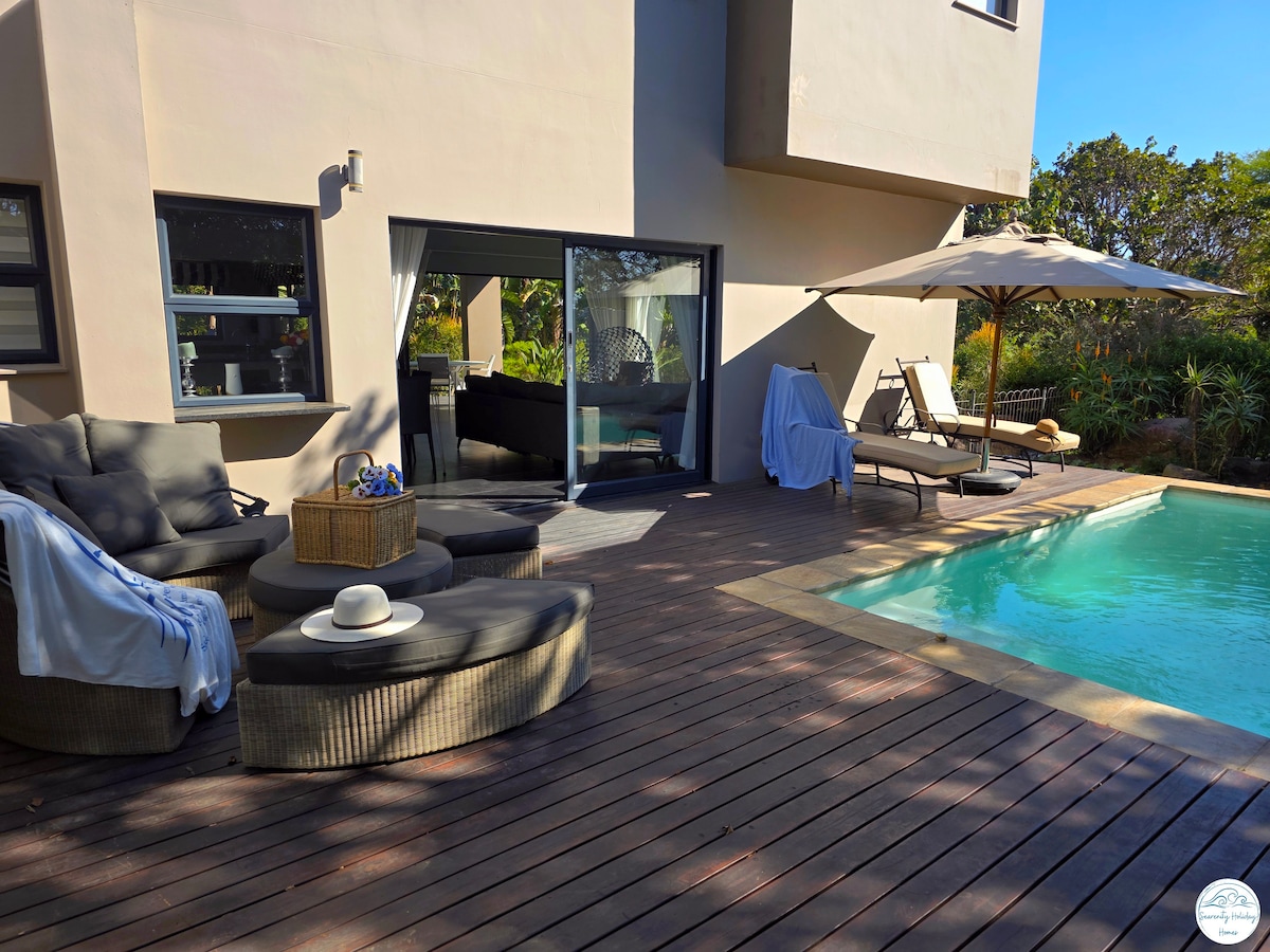 A wooden deck surrounds a sparkling blue pool, equipped with two sun loungers and a shaded umbrella. A circular seating area with cozy, plush cushions is complemented by a small table. Natural greenery is visible in the background, enhancing the outdoor space.