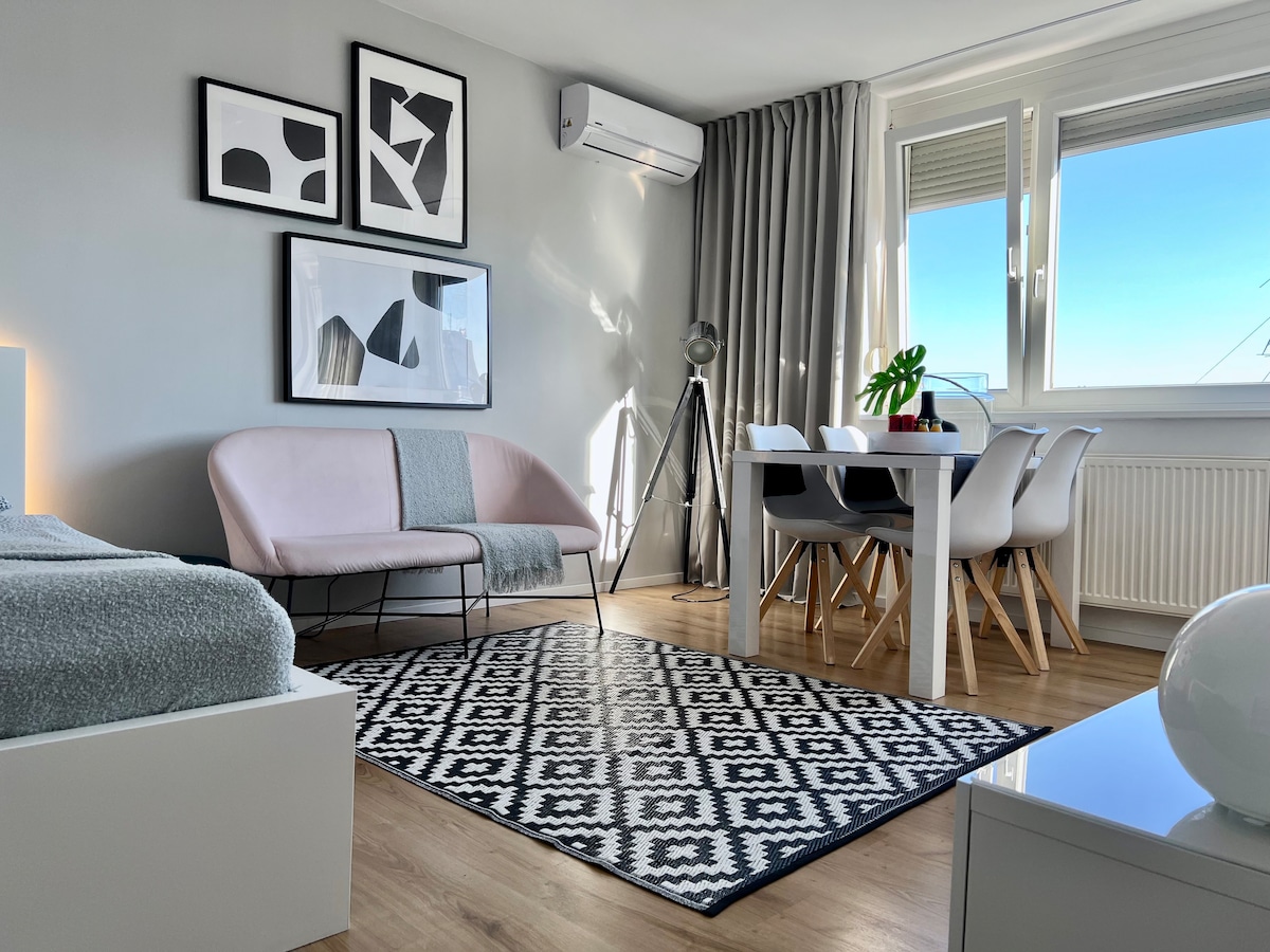 A modern multifunctional living area features a spacious bed with fresh linens, a light pink sofa, and a dining table for four. A patterned area rug anchors the space, with natural light streaming in through large windows, complemented by neutral-toned curtains and stylish wall art.