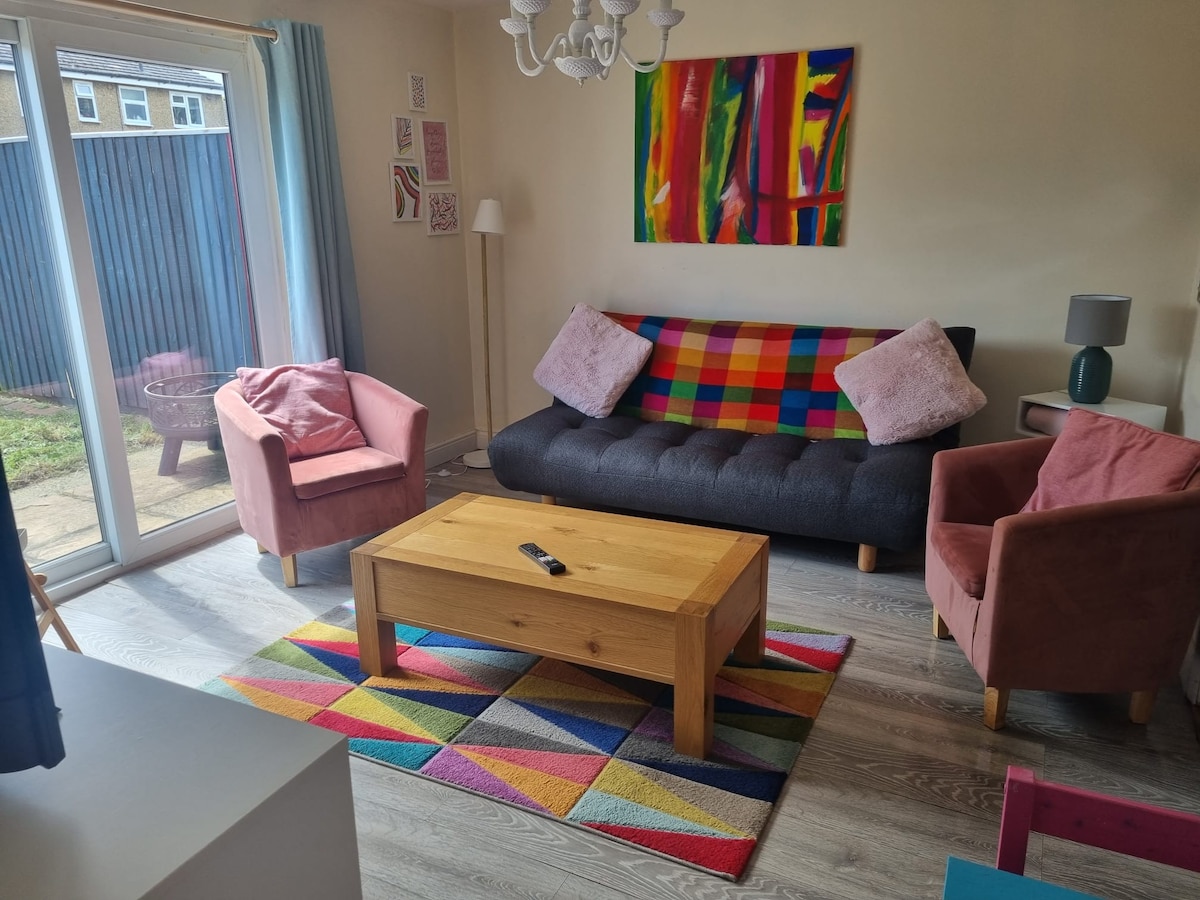 A living room area is presented with a black sofa adorned with colorful throw pillows, paired with two pink armchairs. A wooden coffee table rests on a vibrant, patterned area rug. Natural light enters through sliding glass doors, which lead to a garden space.