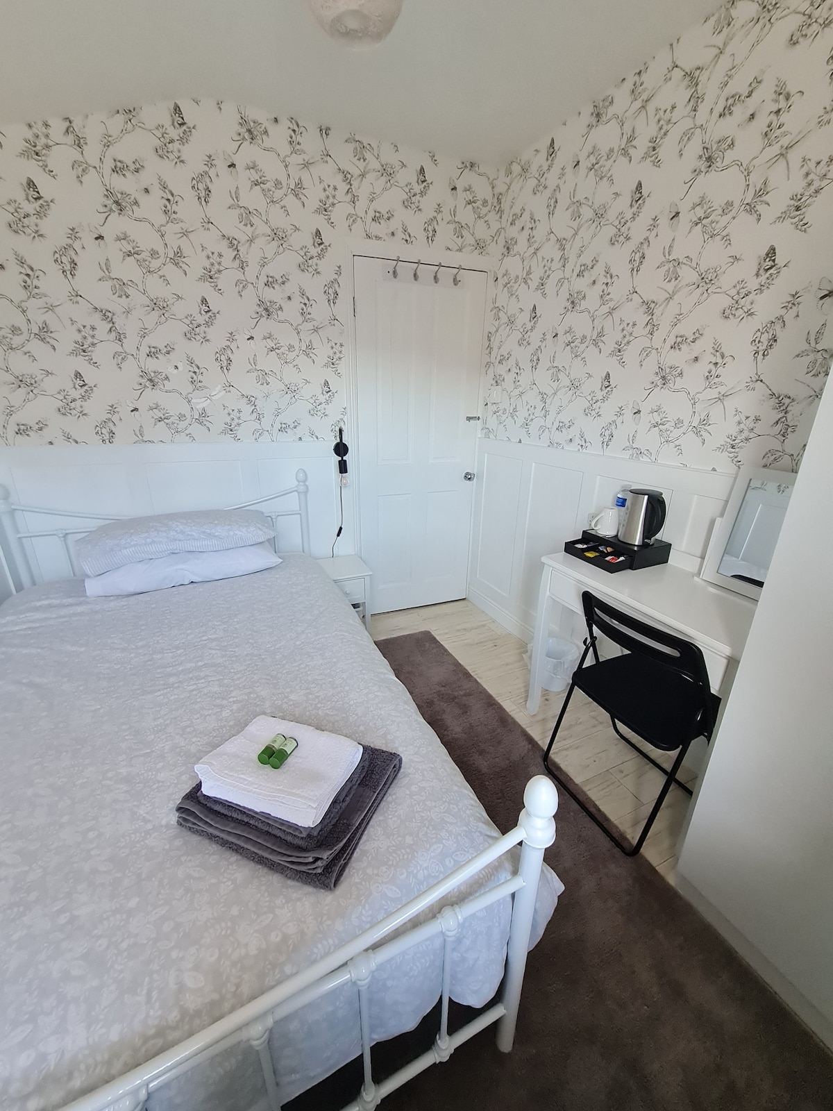 A small bedroom is showcased, featuring a small double bed with fresh linens and neatly arranged towels. A desk with a chair, kettle, and additional amenities is positioned against the wall. Floral wallpaper adds a subtle charm to the serene atmosphere.