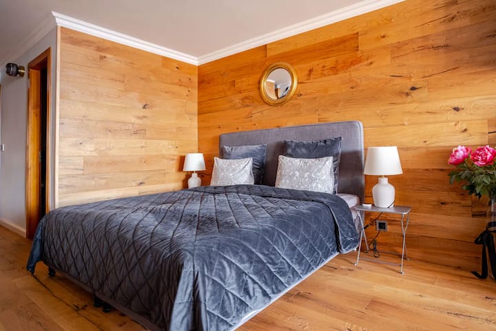 غرفة نوم تحتوي على سرير مزدوج مريح مع وسائد منقوشة وبطانية سوداء مبطنة، محاطة بطاولات بجانب السرير مع مصابيح، وجدار خشبي مزين بمرآة مستديرة.