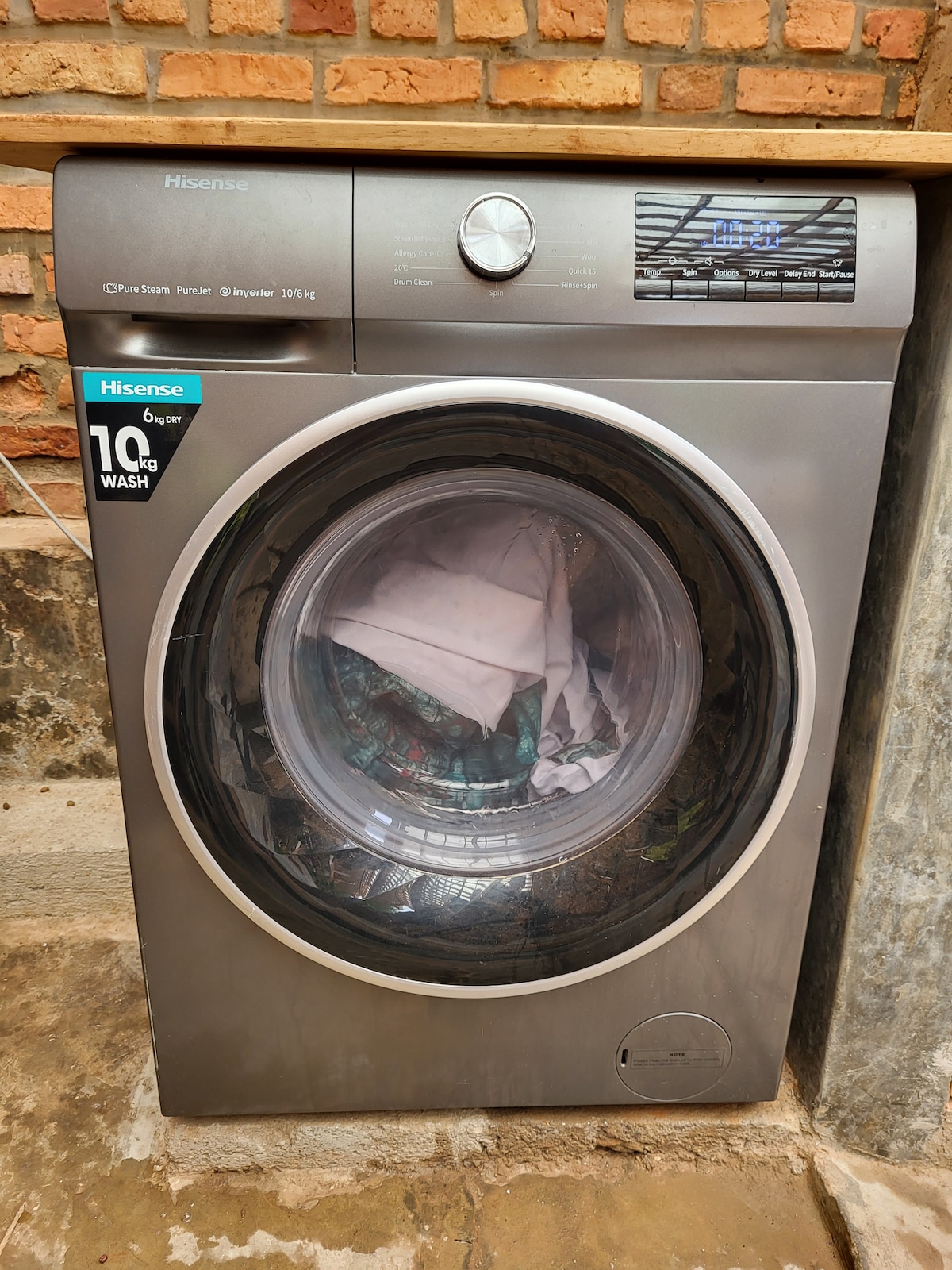 A modern washing machine is displayed with a transparent front door showing a load of laundry inside. The sleek design features a simple control panel and a sticker indicating a 10 kg wash capacity. The machine is set against a textured brick wall.
