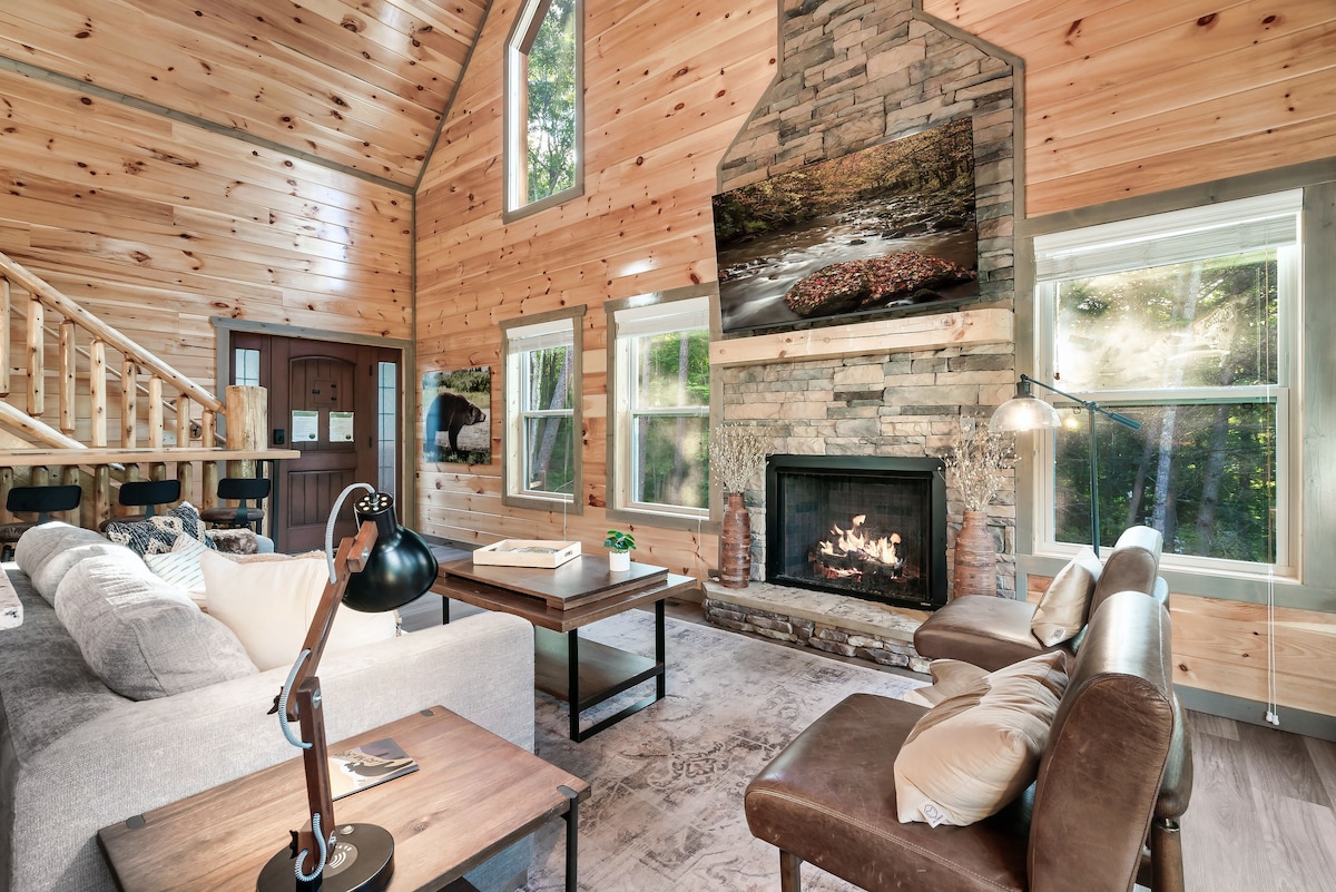 A warmly decorated living area features a stone fireplace with a modern TV mounted above. Large windows provide ample natural light, highlighting the wood paneling and rustic furnishings. A comfortable sofa and armchairs invite relaxation, complemented by a stylish coffee table and floor lamp.