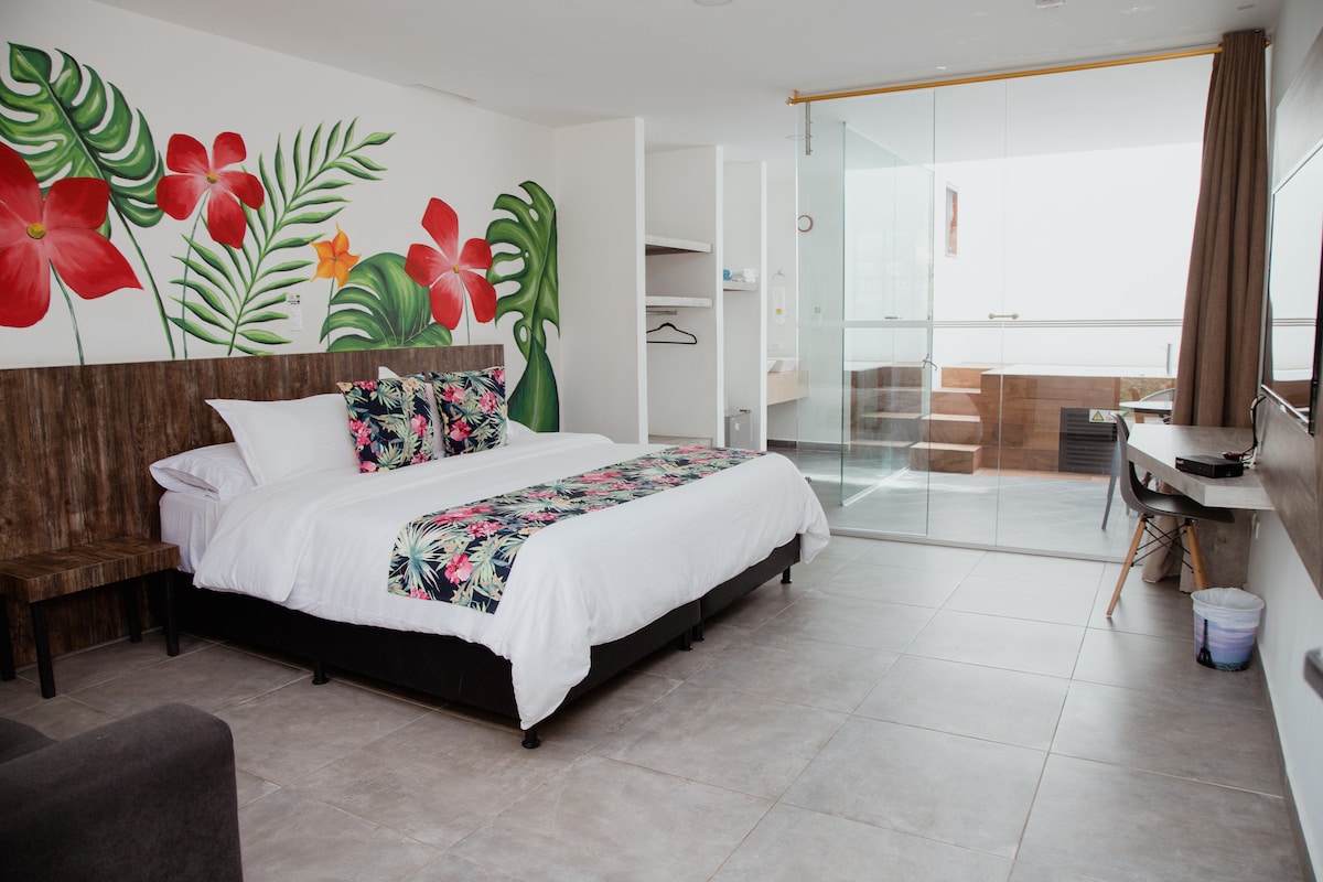 A spacious room features a large bed with a floral-patterned blanket. Vibrant wall art of tropical plants and flowers enhances the decor. A glass door leads to an outdoor area, while natural light fills the room through large windows. A minimalist seating area is present.