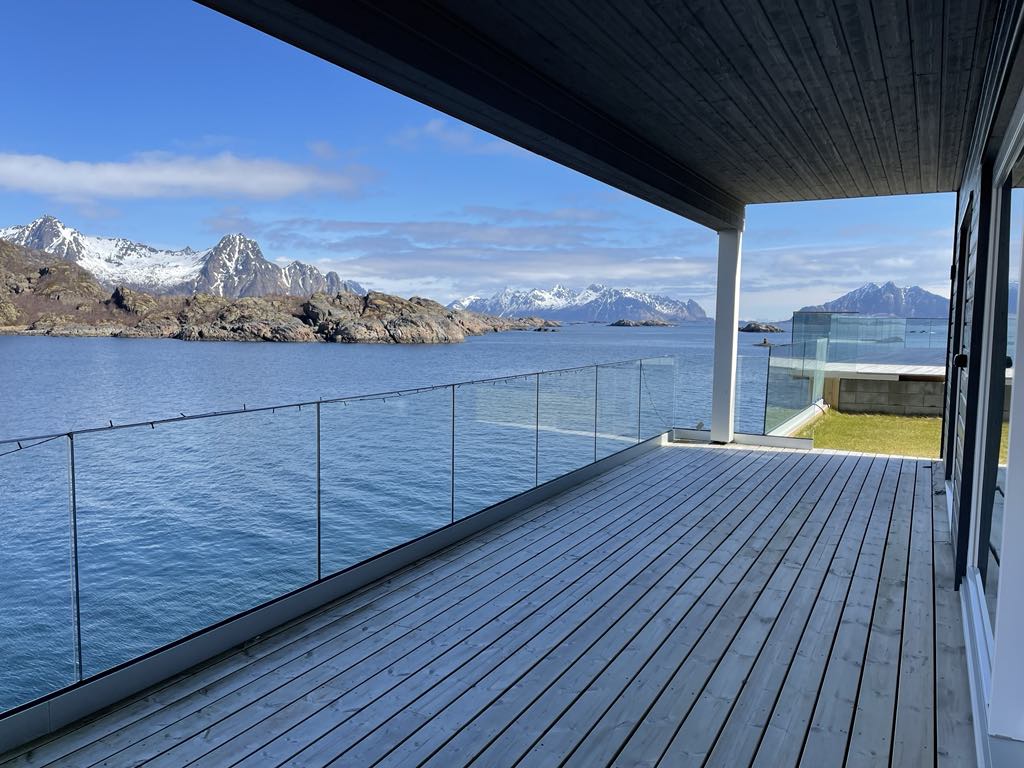 A spacious terrace extends over the water, featuring wooden decking and glass railings that offer unobstructed views of the surrounding mountains and coastline. The landscape reflects a serene atmosphere under a clear blue sky, with gentle waves visible in the foreground.