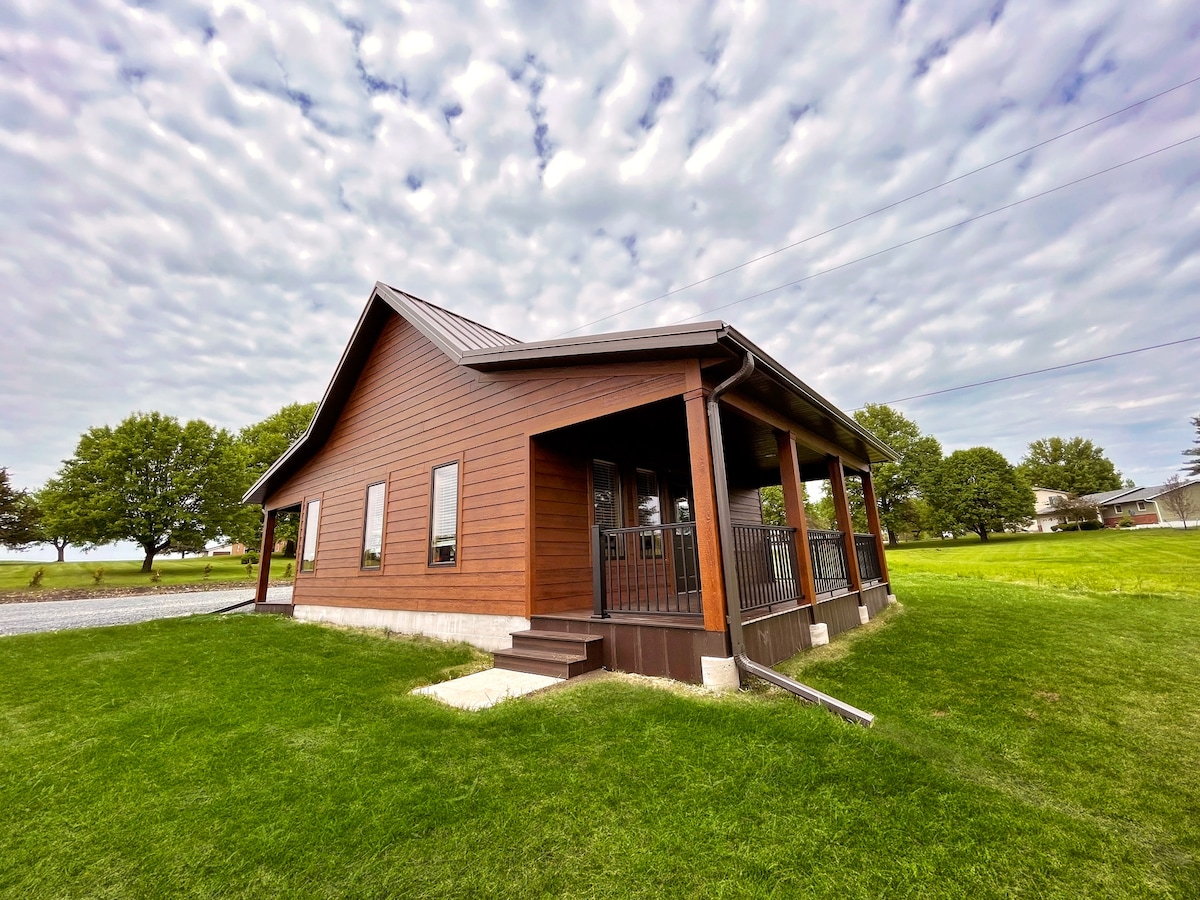A cabin with a wooden exterior is surrounded by lush green grass under a sky scattered with clouds. The structure features a welcoming porch with ample seating options, and large windows allow for natural light to enter the interior.