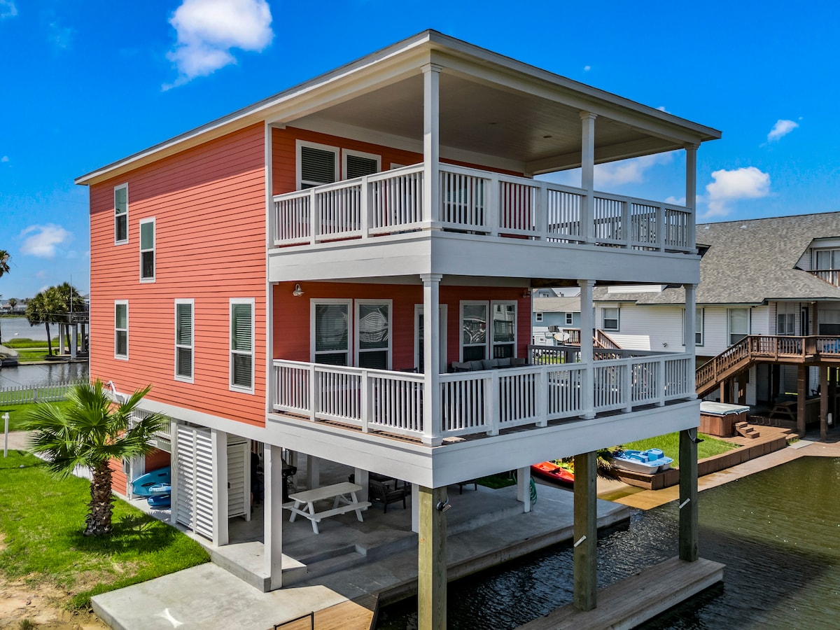 The two-story beach house features a vibrant exterior with large windows that allow ample natural light. A spacious deck is visible on the upper level, offering a relaxing space to enjoy views of the canal. A palm tree adds to the coastal charm in the foreground.