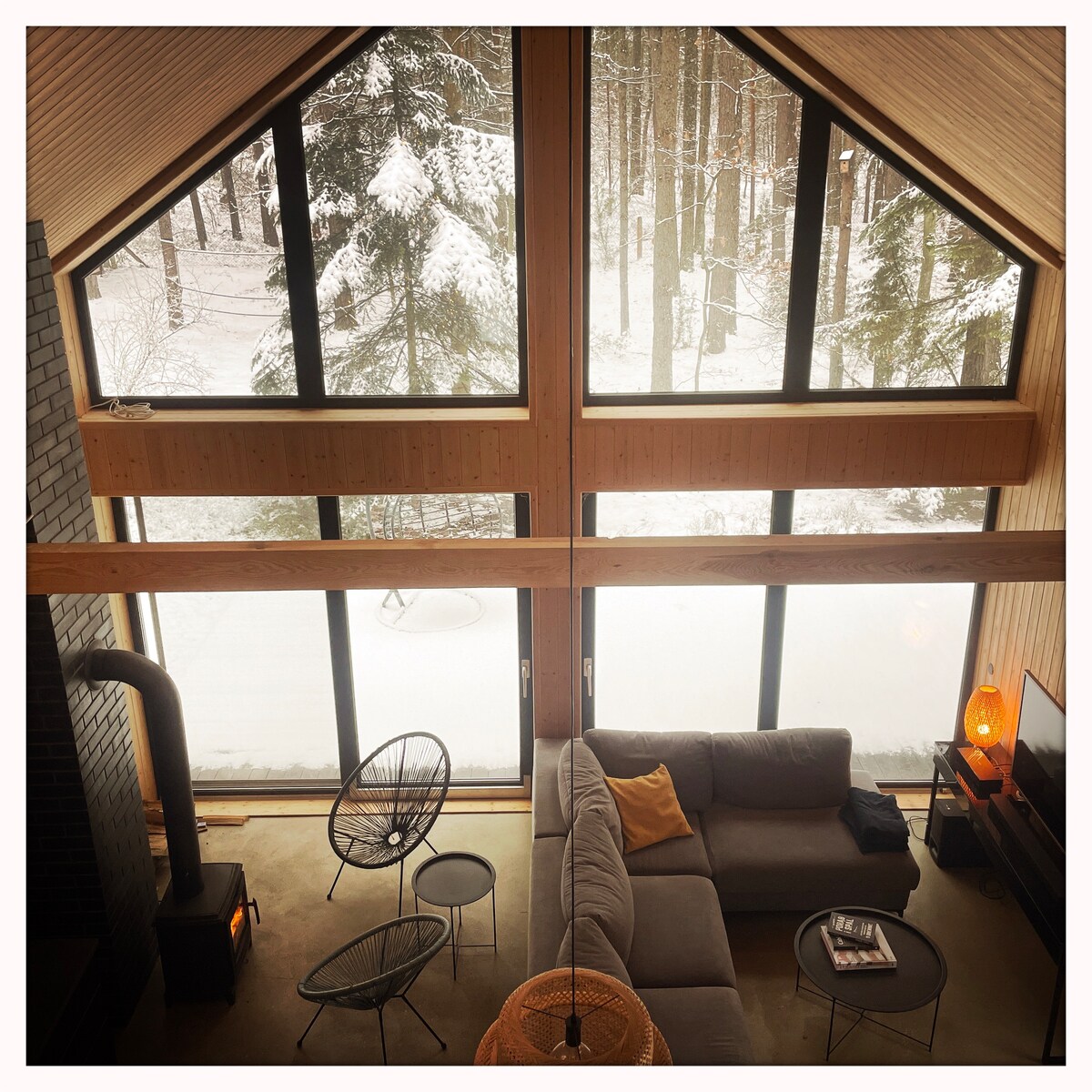 Large windows frame a snowy forest view, allowing ample natural light into the spacious living area. A gray sectional sofa is accompanied by a woven chair and a small coffee table, creating a cozy seating area. A wood stove adds warmth to the space.