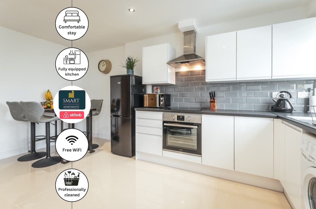 A modern kitchen space features sleek white cabinetry and dark granite countertops. Stainless steel appliances, including an oven and refrigerator, are present. A small dining area with two stools is positioned nearby, and light floods the room through a window, enhancing the spacious feel.