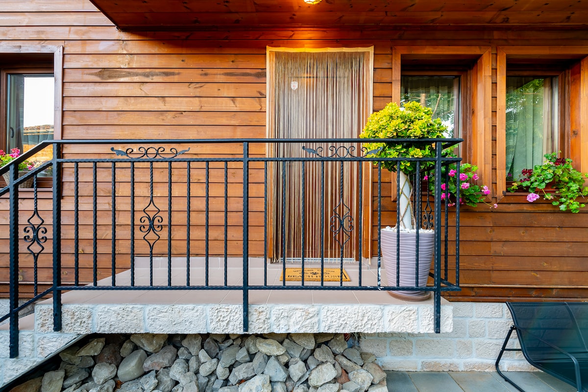 A wooden entrance is framed by a black wrought-iron railing, leading to a welcoming door adorned with a light curtain. Potted plants with colorful blossoms are arranged along the railing, while gravel and stone provide texture to the ground below.