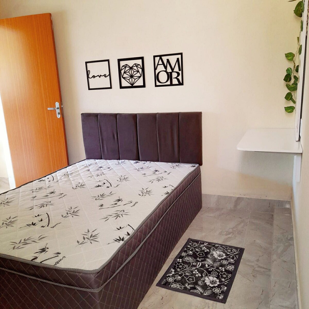 A simple bedroom features a dark upholstered bed with decorative floral bedding. A small wall-mounted shelf is positioned beside the bed, with framed wall art displaying the words 'love' and 'amor.' A patterned rug lies on the tiled floor near the bed.