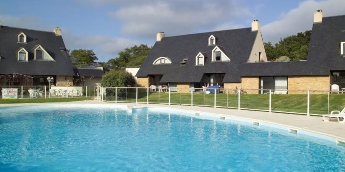 A shared swimming pool is surrounded by a grassy area and low fences. The pool reflects the sky, while several lounge chairs are positioned nearby. Two buildings with pitched roofs can be seen in the background, featuring large windows for natural light.