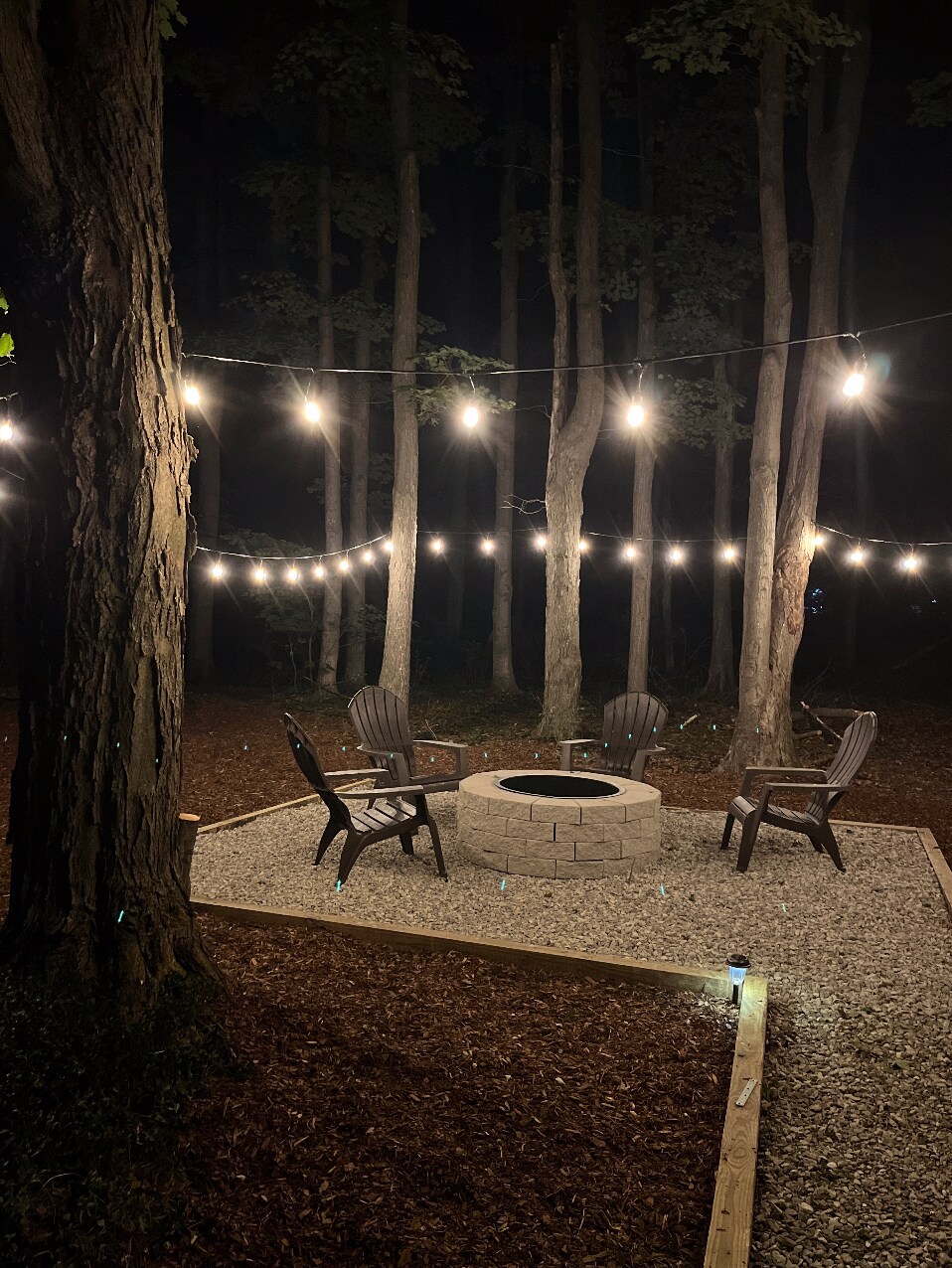 A cozy fire pit area is illuminated by string lights draped between trees. Gravel surrounds the circular stone fire pit, with four Adirondack chairs positioned for comfort. The wooded backdrop enhances the secluded ambiance, creating a perfect spot for evening gatherings.