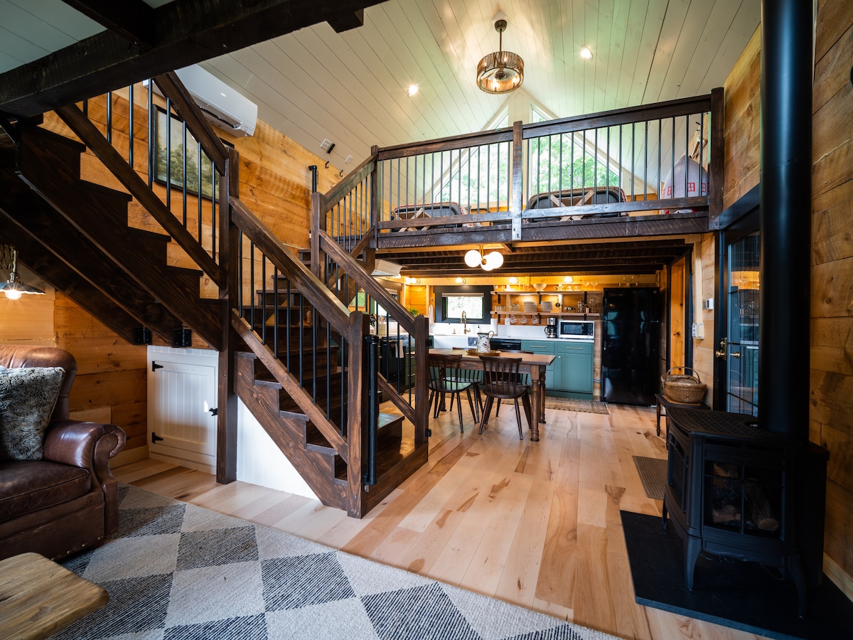The open living area features a wooden staircase leading to an upper loft. A spacious dining table sits next to the modern kitchen, complemented by warm wood tones and cozy furnishings. Soft light illuminates from overhead fixtures, creating a welcoming ambiance throughout the space.