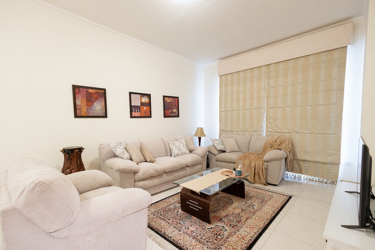 A spacious living room is furnished with light-colored sofas positioned around a coffee table. A textured rug enhances the floor, while framed artwork adorns the walls. Natural light filters through a large window, covered by a beige curtain.