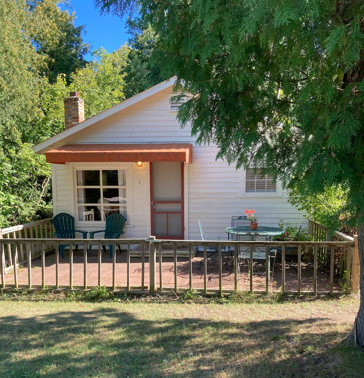 A charming white cottage is framed by lush greenery and a wooden deck. Two blue chairs and a small table with a potted plant are positioned on the porch under a brown awning, inviting relaxation in a peaceful outdoor setting.