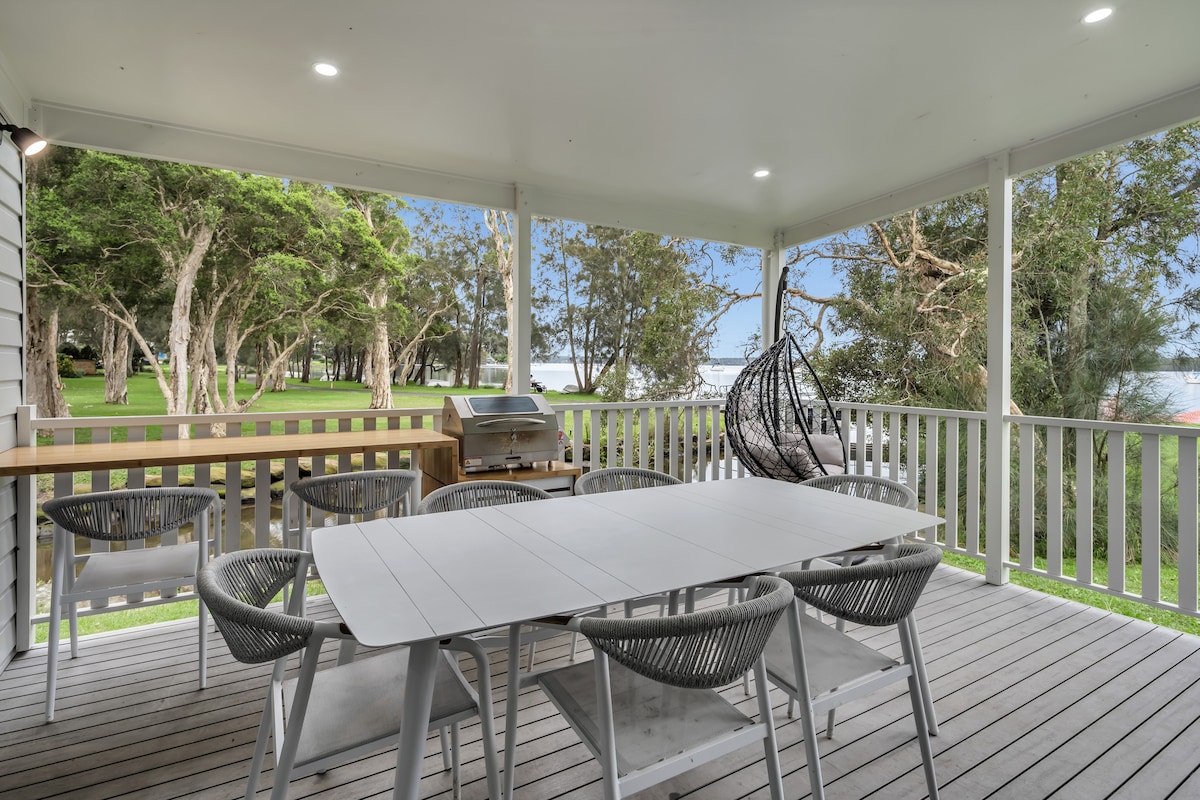 A spacious outdoor deck is featured, equipped with a modern dining table surrounded by several chairs. A grill is visible, complemented by natural views of trees and the waterfront beyond. Soft lighting enhances the space, inviting relaxation and gatherings.