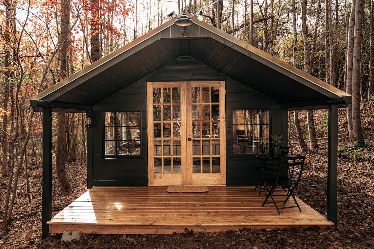 The cabin's exterior features a dark wood façade and a steeply pitched roof, surrounded by tall trees with autumn foliage. Double glass doors provide access to the interior, while a small wooden deck holds a table and chair set, offering a cozy outdoor seating area.