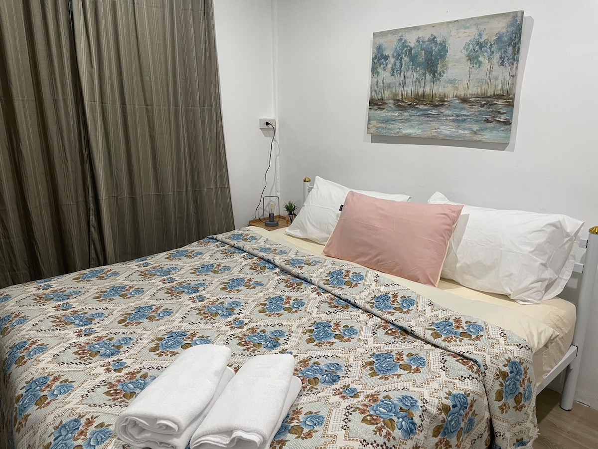 A spacious bed with a floral-patterned blanket is positioned against a soft wall. Two fluffy pillows rest above the bedding. Fresh white towels are neatly arranged at the foot of the bed. A nature-themed painting adorns the wall, complemented by flowing curtains.
