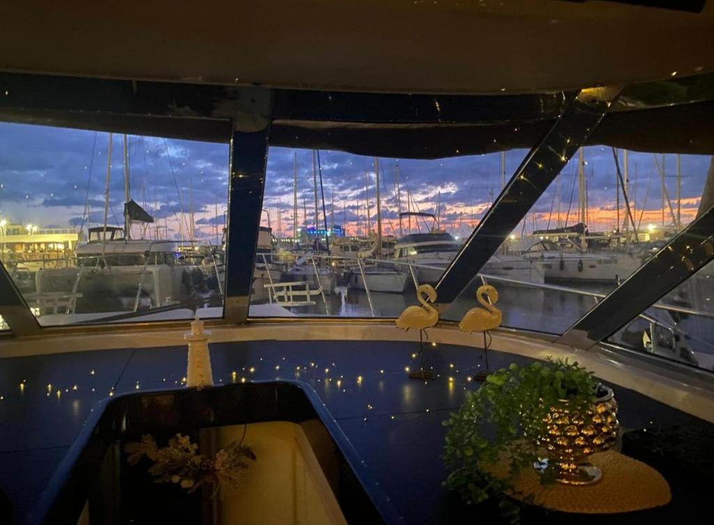 A cozy interior space features large windows that offer views of the marina at sunset. Decorative elements include a plant, a lit candle, and two unique sculptures, enhancing the serene atmosphere. The sky displays a gradient of colors, reflecting the charm of the location.