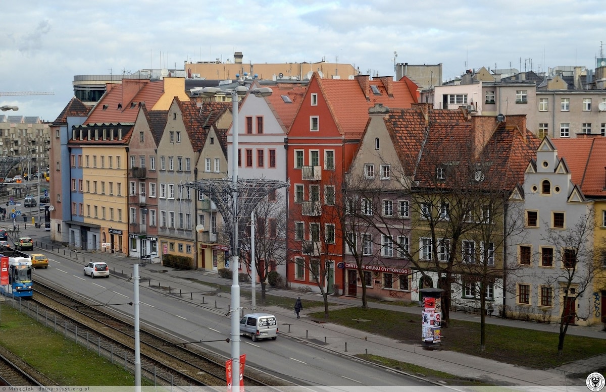 A variety of colorful buildings lines the street, featuring different architectural styles. The structures boast distinct roof designs, with red and orange tiles. Trees are scattered along the sidewalk, and a tram track runs parallel to the street, indicating a lively urban environment.