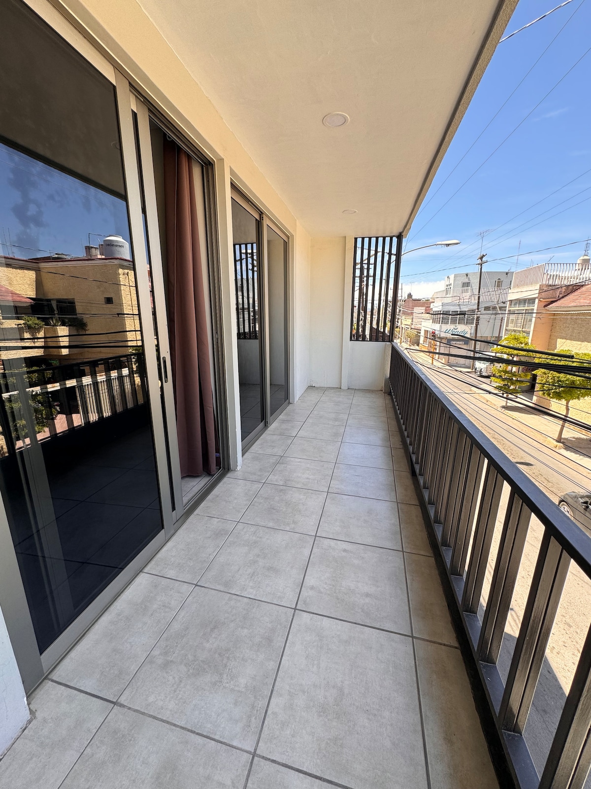 A private balcony is depicted, featuring a tiled surface and glass sliding doors. The railing provides a view of the street below, suggesting a bright and open space. Natural light is visible, enhancing the connection to the outdoors.