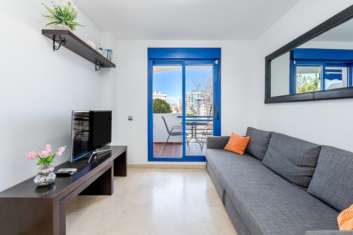 A modern living area features a gray sofa positioned against a white wall, complemented by a black coffee table. A television is mounted on the wall, while natural light enters through a large window showcasing a balcony view. A decorative plant and a small vase with flowers add subtle touches to the space.