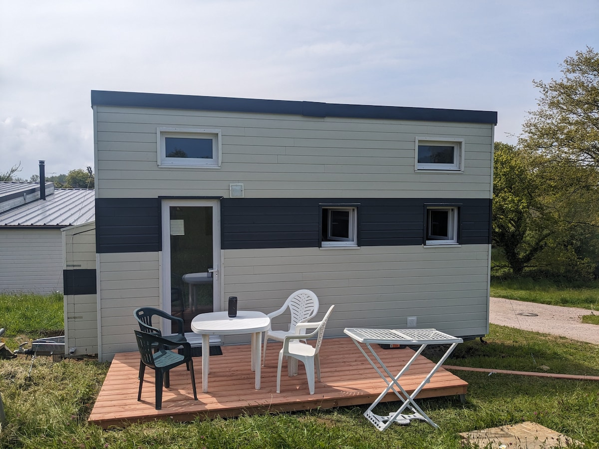 The exterior of the tiny house is framed by green grass, featuring a wooden deck with a white table and four chairs. Large windows allow natural light to fill the space, while the design emphasizes a simple, modern aesthetic.