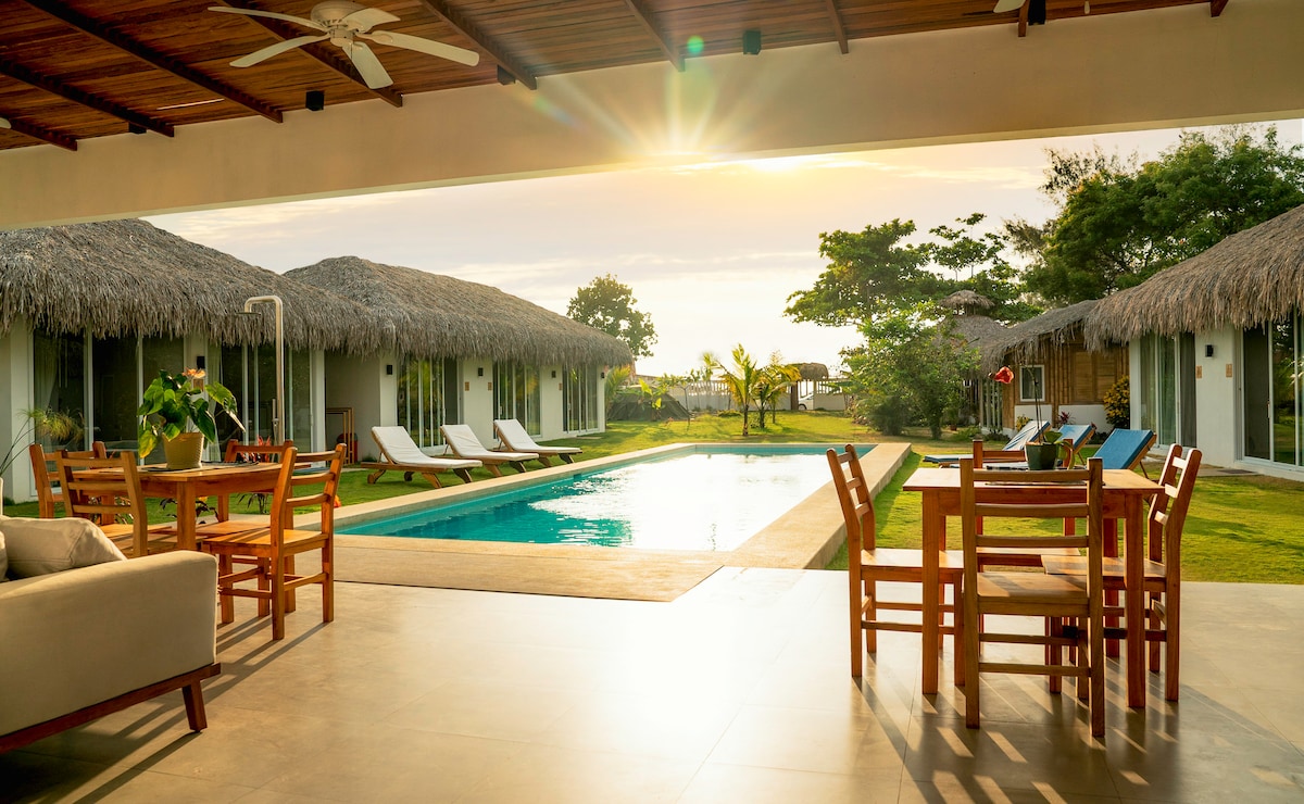A spacious outdoor area is depicted, featuring modern seating arrangements around a central pool. Sunlight streams across the scene, highlighting the lush green garden. A row of loungers is arranged beside the pool, with thatched-roof structures visible in the background.