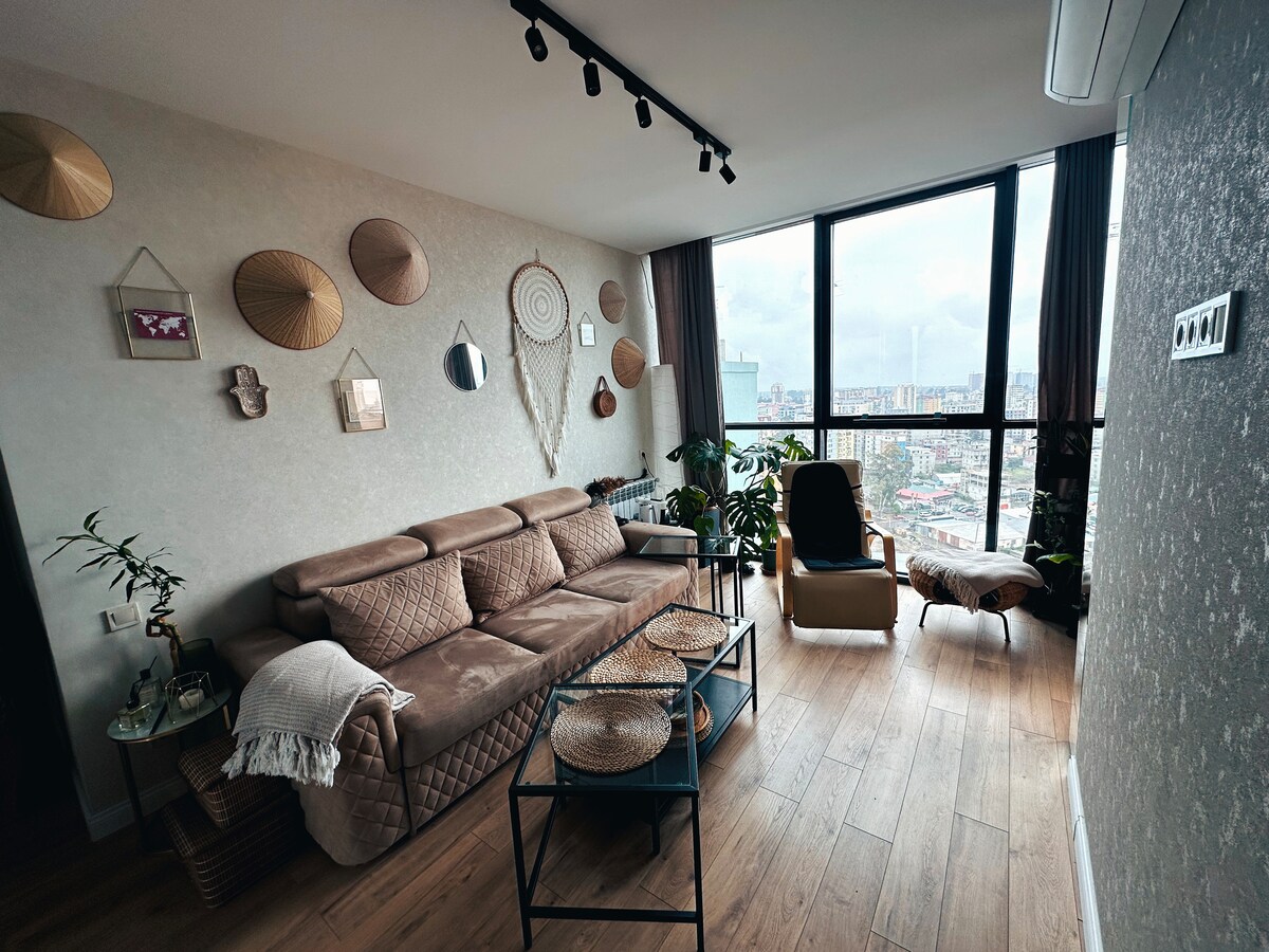 A modern living area features a comfortable sectional sofa complemented by woven wall decor. Large windows provide natural light and a city skyline view. Potted plants add a touch of greenery, while a cozy chair invites relaxation. A coffee table holds decorative trays.