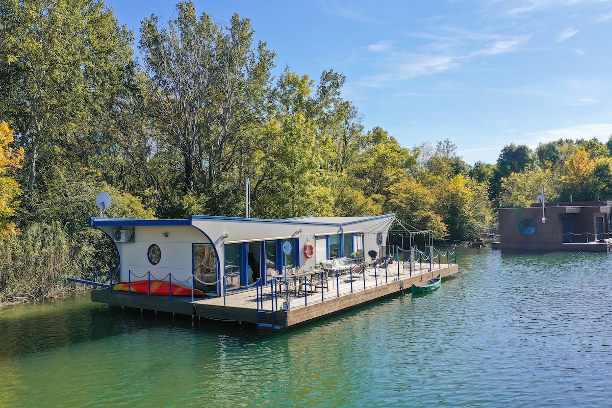 A modern houseboat is anchored in calm water, surrounded by lush greenery. The structure features large windows and a spacious deck equipped with outdoor seating and a kayak. The tranquil setting is enhanced by trees lining the waterfront.