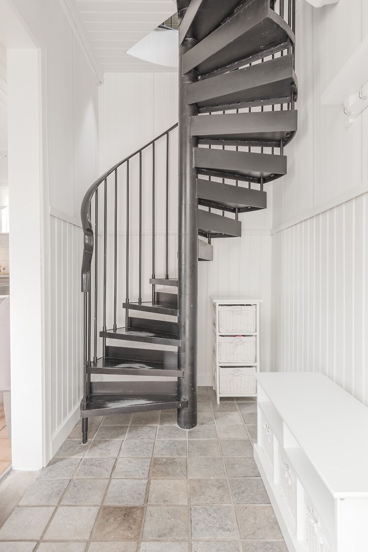 A spiral staircase is positioned against a white panelled wall, showcasing a sleek black metal design. The steps lead upward, while a white storage unit with open compartments stands nearby on a gray tiled floor.