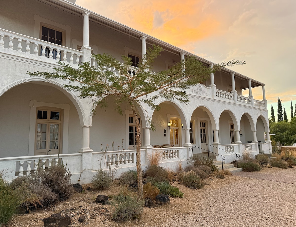 The exterior of a two-story historic building is showcased, featuring a white facade and arched balconies. Lush landscaping with native shrubs and a small tree is visible in the foreground. The sky is lit with warm tones, suggesting sunset lighting.
