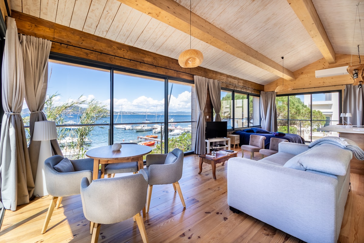 A spacious living area features large glass doors providing unobstructed views of the sea. Natural light fills the room, highlighting the wooden ceiling and flooring. A round dining table and cozy seating arrangement create a comfortable space, while a television and kitchen area are visible in the background.
